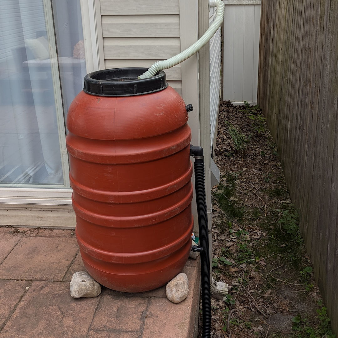 A terra cotta-colored rain barrel on a patio. A flex tube is running from a nearby downspout to the top of the barrel so that rain running off of the roof will be diverted into the barrel.