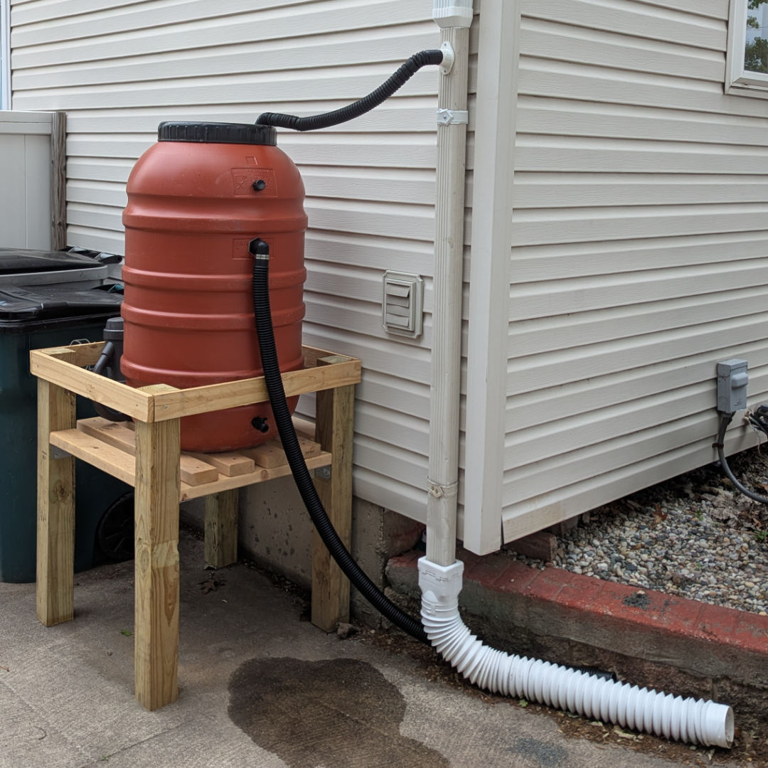 A terra cotta-colored rain barrel, raised up on a wooden stand about 3 feet off the ground, on the other side of the house from where the first barrel is. A flex tube is running from a nearby downspout to the top of the barrel so that rain running off of the roof will be diverted into the barrel.